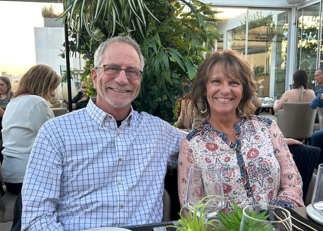 Smiling couple sitting outdoors at a table with greenery.